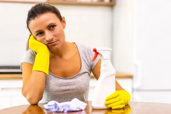 Женщина устала после тяжелой работы по уборке, опираясь на руку в защитной перчатке и держа бутылку спрея на кухонном фоне . — стоковое фото