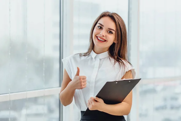 Positive smiling female teacher inspired with education ideas, stands holding paperholder, giving thumb up. — Stock Photo, Image