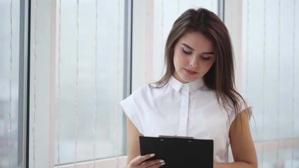 Beautiful businesswoman is taking notes on the clipboard. She is looking at the camera and is smiling widely. Close up. Copy space. 4K. — Stock Video