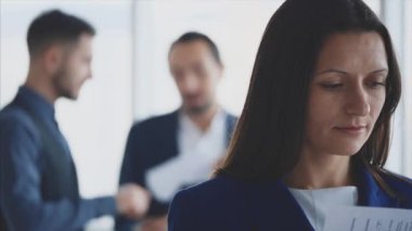 Male business workers are discussing issue on the light blurred background. Woman is turning over pages on the forefront. She is smiling widely. Copy space. Close up. 4K.