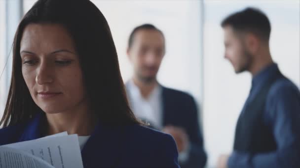 Male business workers are discussing issue on the light blurred background. Woman is turning over pages on the forefront. She is smiling sincerely. Copy space. Close up. 4K. — Stok video