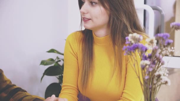 Adorable chica está sentada en la mesa en la cafetería. Ella está hablando con su novio, que sostiene sus manos. El foco cae al florero con las flores y la mesa. De cerca. Copiar espacio . — Vídeos de Stock