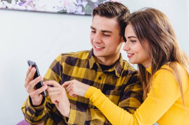 Lively young couple looking through photos on smartphone, smiling with pleasure as they relax at home.