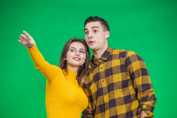 Portrait of young woman pointing at something ahead isolated on green background, her boyfriend is looking at it, pleasantly surprised. — Stockfoto