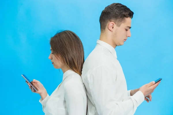 Beautiful young business woman and handsome businessman in formal clothes are using gadgets, standing back to back over blue background. — Stock Photo, Image