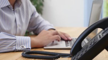 Businessmans hands type at desktop keyboard in workspace. Focus is on the landline phone, blurred background. Close up. Copy space. Crop. 4K.