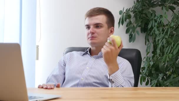 Young businessman is eating apple in the office. Meanwhile he is looking at the laptop. Close up. Copy space. 4K. — Stok video