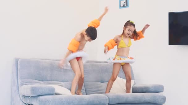 Dos niños pequeños, vestidos con trajes de baño, están felices de ir de viaje. Están bailando en el sofá. Copiar espacio. 4K . — Vídeo de stock