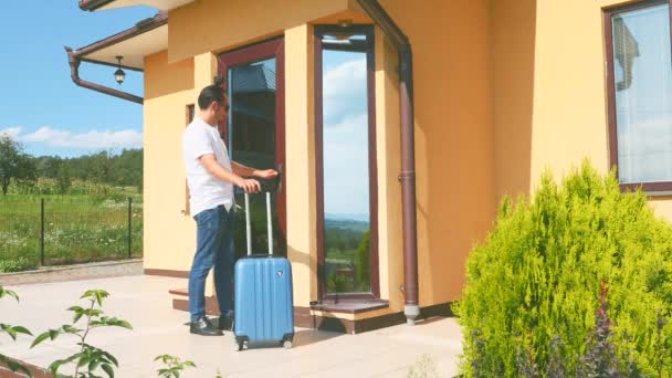 Schöner Mann kommt nach Hause und versucht, die Tür zu öffnen. Aber sie ist geschlossen. Er kommt mit einem blauen Koffer aus dem Urlaub. Leeres Haus. Kopierraum. 4k. — Stockvideo