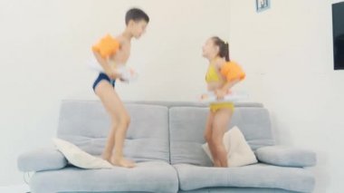 Two little kids, dressed in swimming suits, are happy to go on a trip. They are jumping on the sofa. Copy space. 4K.
