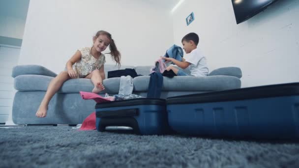 Les petits enfants jettent des vêtements dans la valise à la maison. Ils sont assis sur le canapé bleu. Gros plan 4K . — Video