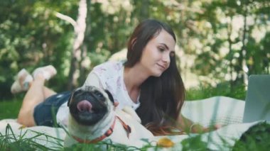 Girl is laying on a carpet on lawn in the park. She typing something on her laptop. Pug laying beside and looking around. Copy space. 4K.