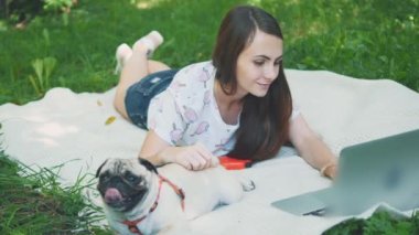 Girl is laying on a carpet on lawn in the park. She typing something on her laptop. Pug laying beside and looking around. Copy space. 4K.