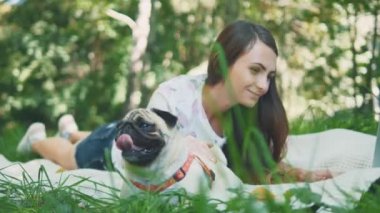 Girl is laying on a carpet on lawn in the park. She typing something on her laptop. Pug laying beside and looking around. Copy space. 4K.