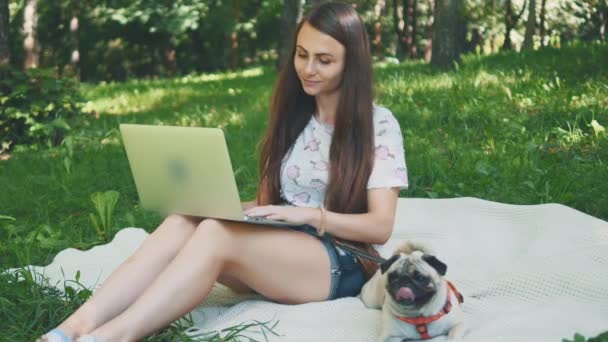 Girl is laying on a carpet on lawn in the park. She typing something on her laptop. Pug laying beside and looking around. Copy space. 4K. — Stock video