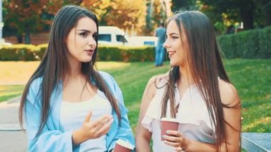 Happy girls friends are talking and smiling holding to-go coffee sitting on bench in park. Action. Movement. 4K.