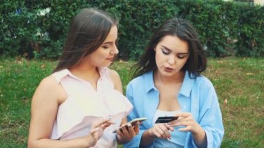 Two attractive girls are shopping online sitting on the bench in the park. Close up. Copy space. 4K.