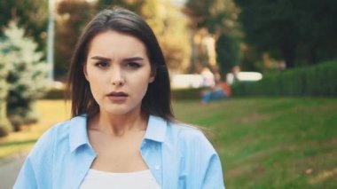 Unhappy girl frawns, being on blurred summer park background. Close up. Copy space. 4K.