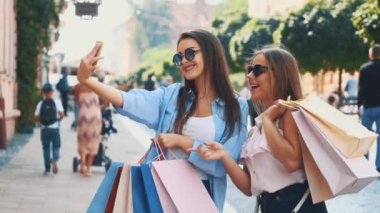 Smiling teenage girls are taking selfie by smartphone, being on summer city park background. They are holding shopping bags. Copy space. Close up. 4K.