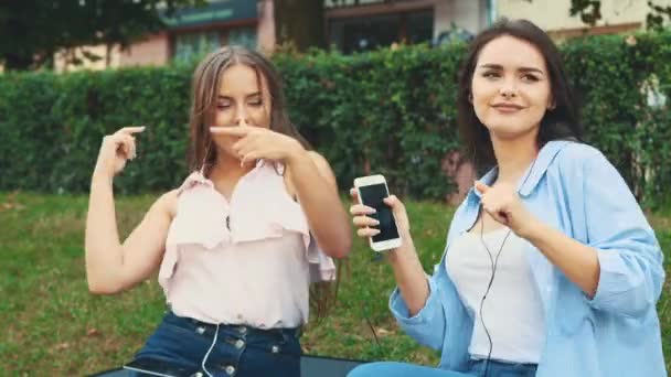 Dos chicas guapas escuchan música usando auriculares en el parque. Copiar espacio. 4K . — Vídeos de Stock