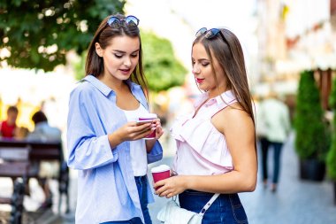 Outdoors fashion portrait of two young beautiful women friends drinking coffee and interactiong after shopping.