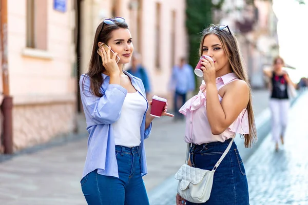 Outdoors fashion portrait of two young beautiful women friends drinking coffee and interactiong after shopping. — Stockfoto