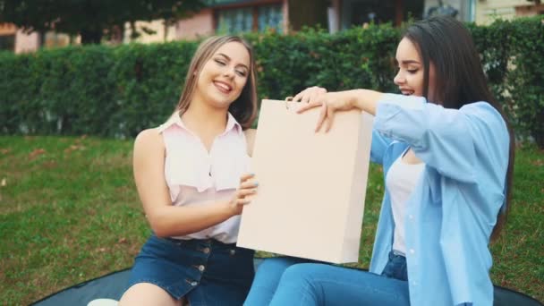 Due ragazze attraenti sono sedute sulla panchina del parco. Ragazza bionda ha comprato una nuova camicetta spogliata per la sua amica bruna. Ricevuto. 4K . — Video Stock