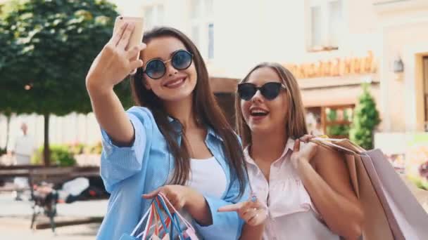 Chicas adolescentes sonrientes están tomando selfie por teléfono inteligente, estar en el fondo del parque de la ciudad de verano. Están sosteniendo bolsas de la compra. Copiar espacio. De cerca. 4K . — Vídeos de Stock