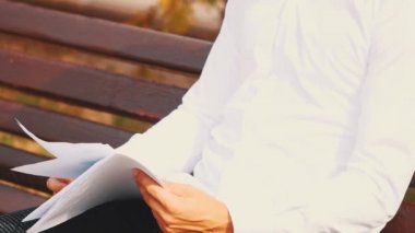 A young unrecognizable businessman is looking through important documents, wearing white shirt, is outside in city, sitting on bench. Crop. Close up. Copy space.
