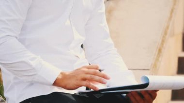 A young unrecognizable businessman with clipboard and pen, wearing white shirt, is outside in city, sitting on bench, writing. Crop. Close up. Copy space.