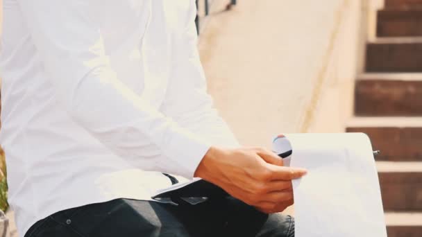 Un joven hombre de negocios irreconocible con portapapeles y bolígrafo, con camisa blanca, está afuera en la ciudad, sentado en el banco, escribiendo. Cultivo. De cerca. Copiar espacio . — Vídeos de Stock