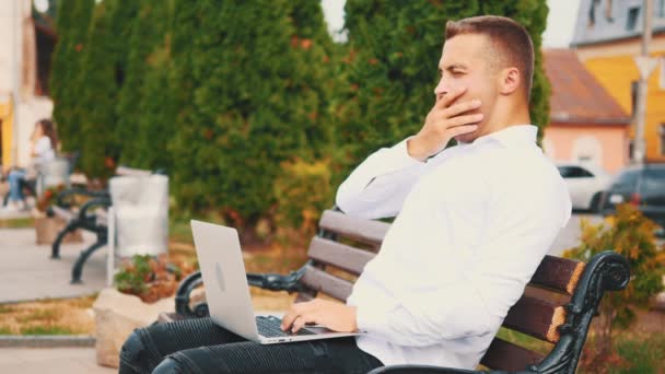 Concentrated man, wearing business style and typing on the laptop computer while sitting outdoors in the park on the bench. Close up. Side view. Copy space. 4K. — ストック動画