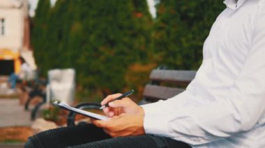 A young unrecognizable businessman with clipboard and pen, wearing white shirt, is outside in city, sitting on bench, writing. Crop. Close up. Side view. Copy space.