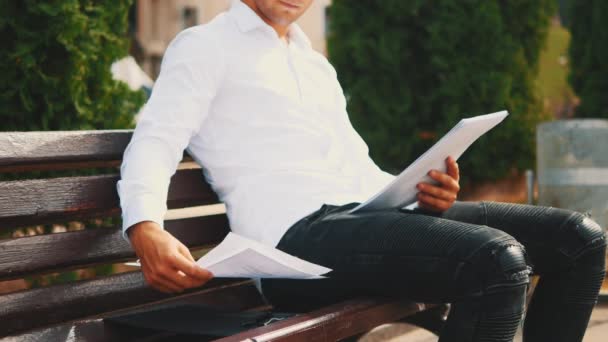 A young unrecognizable businessman is looking through important documents, wearing white shirt, is outside in city, sitting on bench. Crop. Close up. Copy space. — Stockvideo