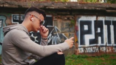 Confident and relaxed guy is dressed in casual clothes. He is listening to cool music with headphones, sitting outside. Close up. Copy space. 4K.