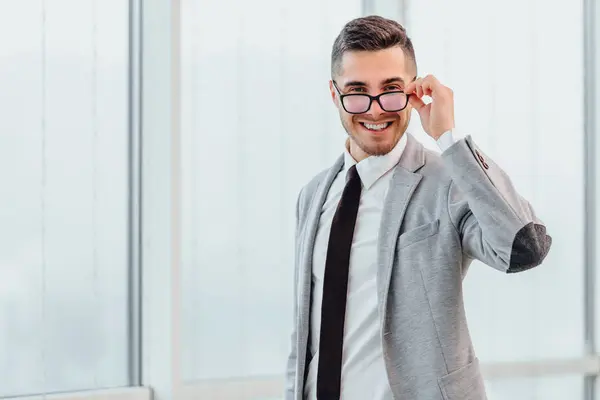 Άνδρας απλός υπάλληλος γραφείου χαμογελά στο γραφείο, στέκεται, αγγίζει τα γυαλιά του, δείχνοντας ενθουσιασμένος. — Φωτογραφία Αρχείου