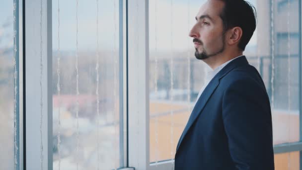 Empresario en suite formal se encuentra frente a la ventana panorámica con vista a la ciudad. Un hombre serio. Vista de perfil. El hombre cruza los brazos. De cerca. Animación. 4K . — Vídeo de stock