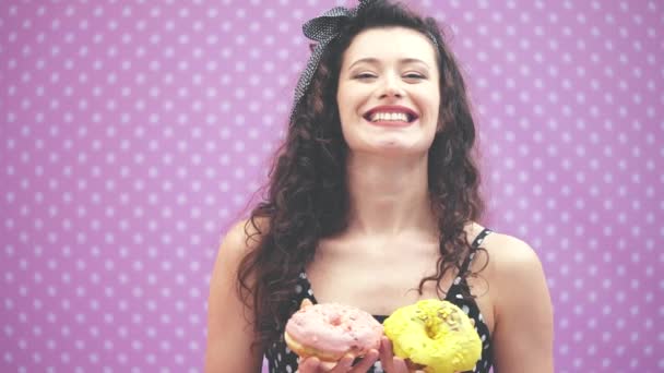 Lovely young curly girl standing with two yummy doughnuts in pink and yellow icing in her hands, grinning happily, showing her satisfaction. — Stock Video