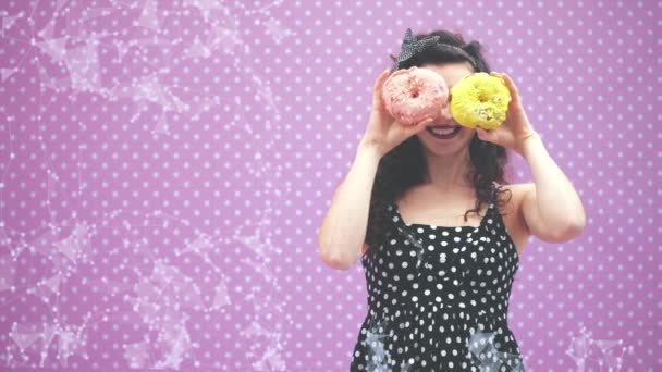 Heerlijk jong krullend meisje verbergt haar ogen achter twee lekkere donuts in roze en gele glazuur, vrolijk grijnzend. — Stockvideo