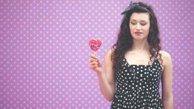 Güzel genç kıvırcık kız ayakta duruyor, elinde çubuğun üzerinde tatlı kırmızı bir lolipop tutuyor, karnını okşuyor, şekere bakıyor,.