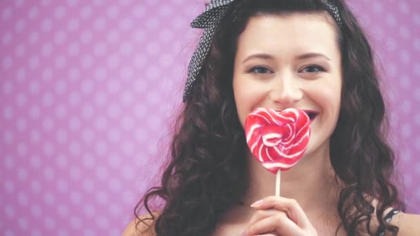 Krásná mladá kudrnatá dívka skrývající ústa za červeným lízátkem na tyčince.. — Stock video