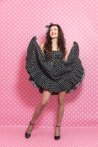 Volledige lengte van pin-up krullend meisje het verhogen van haar polka-dot zwarte jurk, omhoog kijken, poseren, glimlachen mooi. — Stockfoto
