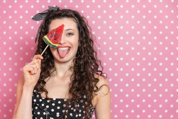 Jeune femme drôle portant pin-up et robe à pois, jouant autour, montrant la langue et l'oeil caché derrière bonbons en forme de pastèque . — Photo