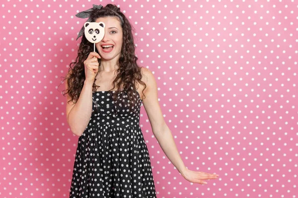 Adorável jovem garota encaracolado com sorriso doce está escondendo seu olho atrás do pirulito na vara, que se parece com panda . — Fotografia de Stock