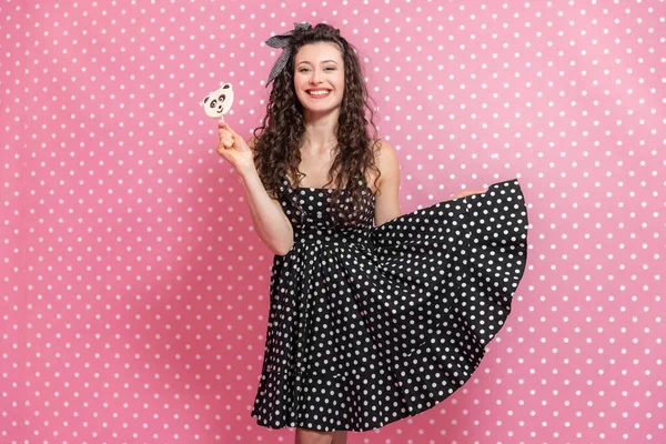 Expressive young female is holding a lollipop on the stick and raising one side of her dress, smiling lightly to the camera. — Stock Photo, Image