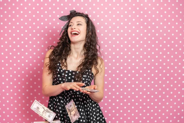 Χαρούμενη νεαρή γυναίκα πετάει χαρτονομίσματα δολαρίων από την παλάμη της ένα προς ένα και γελάει απελπισμένα, υψώνοντας το πηγούνι της και κοιτώντας ψηλά.. — Φωτογραφία Αρχείου