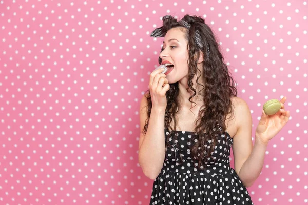 Curly young lady having a snack, tasting delicious macaroon, looking to the side wiith copyspace for remark or product.