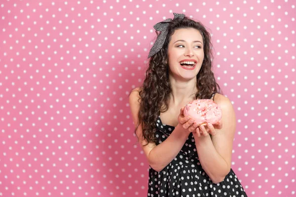 Jovem alegre no vestido pontilhado fica com saboroso donut em um fundo rosa . — Fotografia de Stock