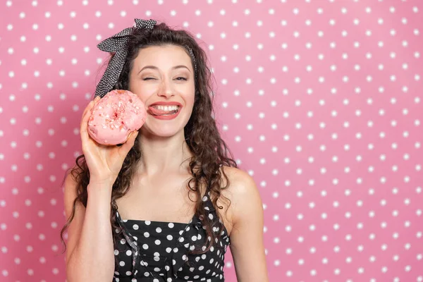 Glamouröses junges Mädchen hält leckeren Donut in der Hand, streckt ihre Zunge heraus und versucht, ein rosa Zuckerguss zu lecken und zwinkert in die Kamera. — Stockfoto