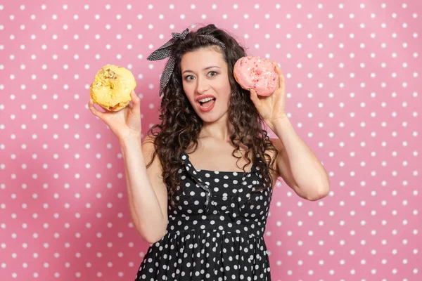 Attraktives Weibchen prahlt mit zwei großen Donuts mit gelbem und rosa Zuckerguss und blickt mit unerklärlichem Gesichtsausdruck in die Kamera. — Stockfoto